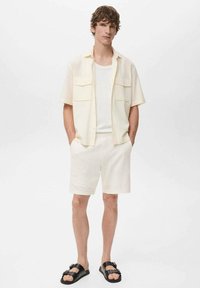 Chemise à manches courtes, de couleur beige clair, avec deux poches sur la poitrine, associée à un short texturé blanc cassé et des sandales noires. Design simple et décontracté.