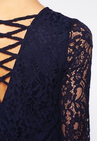 Marinblå spetsoberol med v-ringning, långa ärmar med blommönster och en korslagd detalj på baksidan. Strukturerad tyg med genomskinliga inslag.