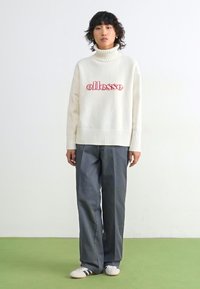Hvit, strikket høyhalset genser med rødt "ellesse"-logo; parret med grå vide bukser og hvite sko med svarte striper.