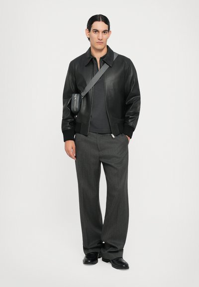 Veste bomber en cuir noir avec des poignets côtelés, chemise en maille grise, pantalon rayé gris ample et chaussures noires ; petit sac bandoulière.