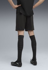 Cilvēks stāv ar rokām uz gurniem, apģērbts melnās futbolā īsās biksēs, ar ceļgaliem augstiem melniem zeķēm un melnām Puma zābakiem uz pelēka fona.