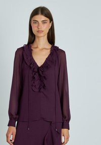 Blusa viola con scollo a V profondo, dettagli a volant lungo il collo e maniche lunghe trasparenti. Presenta lacci con coulisse nella parte inferiore.