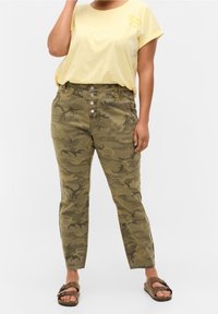 Olivové maskáčové kalhoty s vysokým pasem, pěti knoflíky a bočními kapsami, doplněné žlutým tričkem s grafickým potiskem. Hnědé sandály.