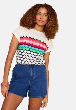 Kvinde med krøllet hår iført en t-shirt med flerfarvet bølget mønster, blå denimshorts, lyserøde øreringe og et lyserødt armbånd.