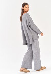 Szary, przytulny sweter z dzianiny o szerokim kroju, zestawiony z pasującymi spodniami o szerokich nogawkach. Modelka nosi beżowe buty typu slip-on.