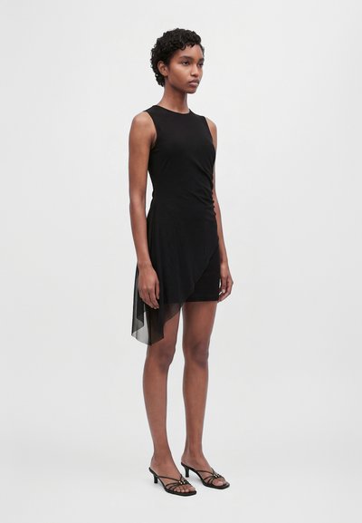 Un mannequin se tient devant un fond uni et clair, vêtu d’une robe noire sans manches avec un voile transparent asymétrique et de chaussures noires à talons avec des brides.