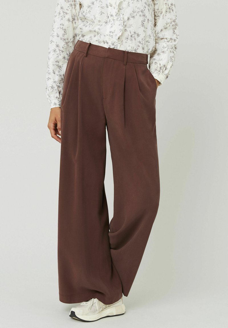 Pantalons marron à jambes larges avec plis à l'avant, présentant une texture lisse. Associés à un blouse à fleurs et des baskets blanches.