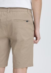 Beige shorts met een elastische tailleband, twee achterzakken en een klein logo-label boven de rechterzak. Glad oppervlak en knielange snit.