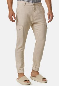 Pantalones cargo de lino beige con tobillos angostos, bolsillos laterales y cierre de botón. Llevados con sandalias beige tipo slip-on. Camisa a rayas visible arriba.