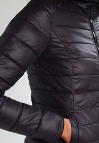 Chaqueta tipo puffer negra con acolchado horizontal, material ligero y cierre con cremallera. Cuenta con bolsillos laterales y una textura suave y brillante.