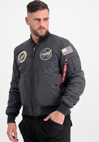 Giubbotto bomber nero con marchio NASA, caratterizzato da polsini e orlo a coste, con toppe ricamate, una tasca con zip e un'etichetta di stoffa rossa sulla manica.