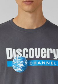 Grijze katoenen t-shirt met een grote blauwe aarde-afbeelding, "Discovery" in vetgedrukte witte tekst, en "CHANNEL" in blauw daaronder.