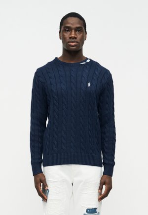 Homme portant un pull en maille torsadée bleu marine avec petit logo blanc et jean blanc déchiré, debout devant un fond clair uni.