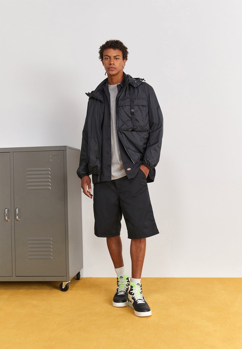 Giacca nera impermeabile con cappuccio regolabile, t-shirt grigia, pantaloni corti neri e sneakers nere con dettagli verde neon. Armadietto grigio sullo sfondo.