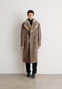 Abrigo marrón de doble botonadura con un gran collar de piel, bolsillos laterales y mangas largas, usado sobre un suéter de cuello alto negro y pantalones negros.