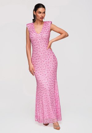 Femme posant dans une robe rose sans manches, longue jusqu'au sol, ornée de sequins avec un décolleté en V et un motif floral, portant des sandales argentées à talons hauts.