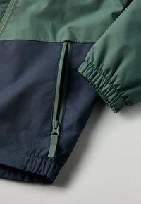 Groene en marineblauwe jas met een ritszak, elastische manchetten en een gladde, lichte textuur, ontworpen voor comfort en functionaliteit.