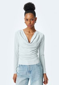 Hellblaues Langarmoberteil mit drapierter Halslinie und gerafften Stoffdetails, kombiniert mit hochtaillierten hellblauen Jeans.
