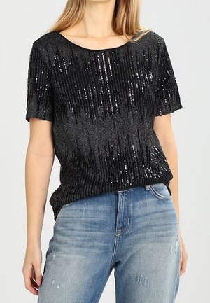 Kobieta w krótkim, czarnym topie z cekinami w pionowe paski, w połączeniu z jasnoniebieskimi, podniszczonymi dżinsami.