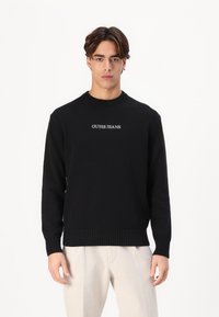 Fekete kötött pulóver, bordás környakú és díszített mandzsettákkal, fehér "GUESS JEANS" hímzéssel a mellrészen.