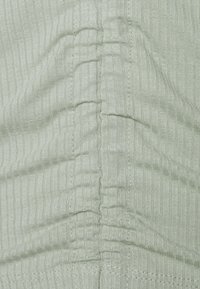 Heller grüner, gerippter Stoff mit vertikalen Texturlinien und gerafften Nähten, die ein dezentes Wellenmuster auf der Oberfläche erzeugen.