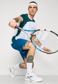 Teniszjátékos zöld és fehér pólóban, kék rövidnadrágban, ütőt tartva. Tetoválásokkal, fehér sportcipőkkel és csuklópántokkal.