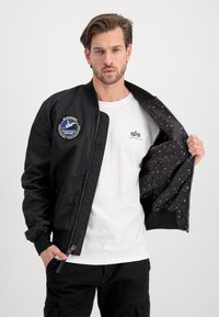 Black bomber jacket with a NASA-inspired patch, zip closure, and ribbed cuffs, featuring a star-patterned lining. Worn over a white T-shirt.