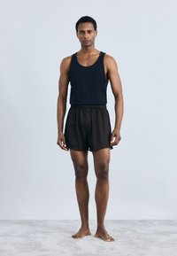 Must mustriga mustriga top, mida kannab mudel koos lõdva musta lühikeste pükstega, millel on peen tekstuur. Mudel seisab paljajalu heledal hallil pinnal.