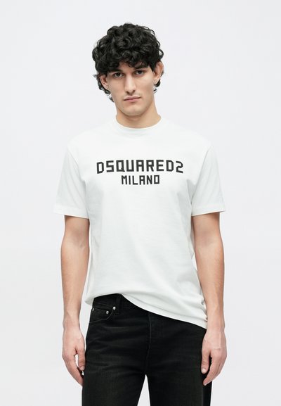 Nooremees lokkis juustega, kes kannab valget "DSQUARED2 MILANO" T-särki ja musti pükse, taustal on lihtne hele toon.