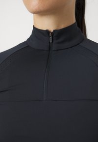 Svart glidelås langermet topp med høy krage, laget av glatt, stretchy materiale. Har perforerte detaljer langs skuldrene.