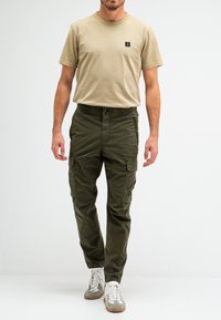 Beige t-shirt met korte mouwen en een klein zwart logo, gecombineerd met olijfgroene cargobroek met zijzakken en een relaxte pasvorm. Witte sneakers.