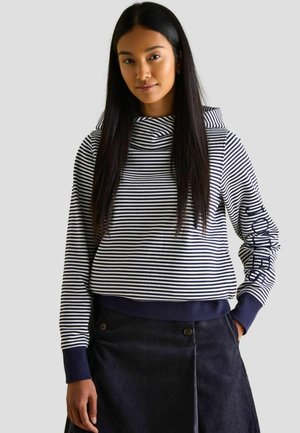 Gestreifter Pullover mit einem Schalkragen, marineblauen und weißen Streifen, gerippten Bündchen und einem dunkelblauen Bund, kombiniert mit einem dunklen Cordrock.