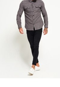 Camisa de button-up gris con dos bolsillos en el pecho, combinada con pantalones slim-fit negros y zapatillas blancas, exhibida en un modelo caminando.