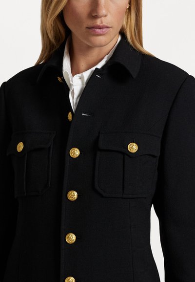 Veste en laine noire dotée d'un col, de boutons dorés et de deux poches avant. Tissu texturé avec une coupe ajustée et des détails de couture visibles.