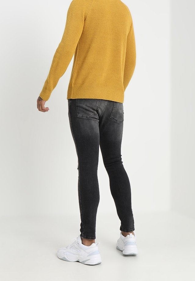 Personne portant un pull moutarde, un jean skinny noir délavé et des baskets blanches, debout de dos sur un fond blanc.