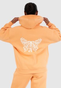 Oranje hoodie gemaakt van zachte stof, met een grote witte vlinderafbeelding op de achterkant en een capuchon met trekkoord. Gladde textuur en een ontspannen pasvorm.