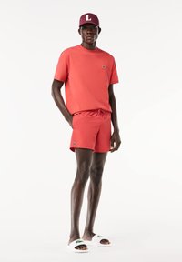 T-shirt rossa in cotone e pantaloni corti coordinati con vita elastica. Il modello indossa ciabatte bianche con logo verde e un cappellino bordeaux con una "L."