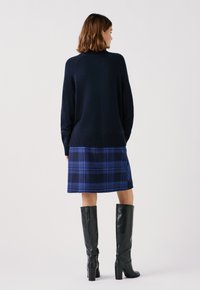 Maglione blu navy a maglia con orlo a costine, abbinato a una gonna scozzese blu con tonalità più scure e stivali neri in pelle al ginocchio.