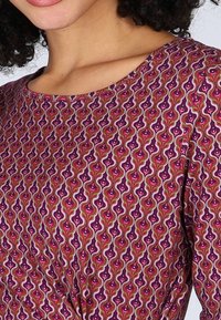 Musterstoff in Lila mit wirbelnden grünen und rosa Motiven, ergänzt durch kleine weiße Akzente. Rundhalsausschnitt, glatte Textur.