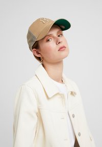 Beige baseball cap med en grøn underside, prydet med et hævet, broderet logo. Meshsider og en solid skygge. Hvid snorjakke tekstur.