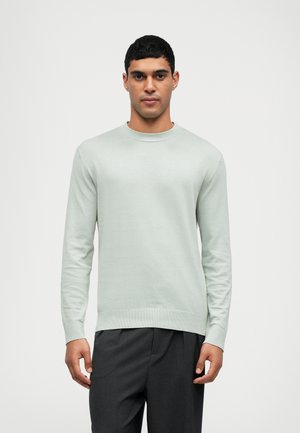 Lys grøn strikket sweater med rund hals og lange ærmer, der har ribbet kant og manchetter, parret med mørkegrå bukser.