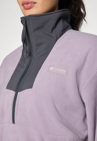 Fialový fleece pulóver s šedým vysokým golierom. Obsahuje zips a nášivku s logom Columbia na hrudi. Mäkka textura a dizajn v blokových farbách.