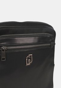 Borsa in pelle nera con hardware argentato, dotata di tasca frontale con zip e una superficie liscia. Dettaglio del logo visibile sulla superficie.