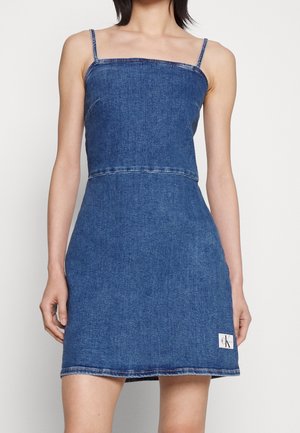 Woman wearing a sleeveless blue denim dress with thin straps and a small logo patch near the hem on the left side.