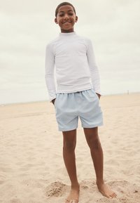 Vit långärmad rashguard och ljusblå badshorts. Strandmiljö med sandig mark och mulet väder. Enkel design, ledig passform.