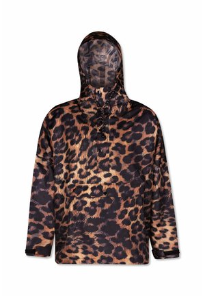 Vrouw draagt een capuchonjas met luipaardprint, donkerbruine broek met boorden, witte sokken en zwarte loafers, staand tegen een effen grijze achtergrond.