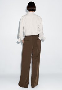 Bílá košile s dlouhými rukávy a knoflíkovými manžetami, doplněná o hnědé kalhoty s vysokým pasem a širokými nohavicemi. Hladká látka se subtílními zadními kapsami.
