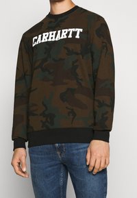 Bruin-groen camouflage sweatshirt met een wit "CARHARTT" logo, ronde hals en zwarte geribbelde boorden aan mouwen en zoom. Zachte stoftextuur.