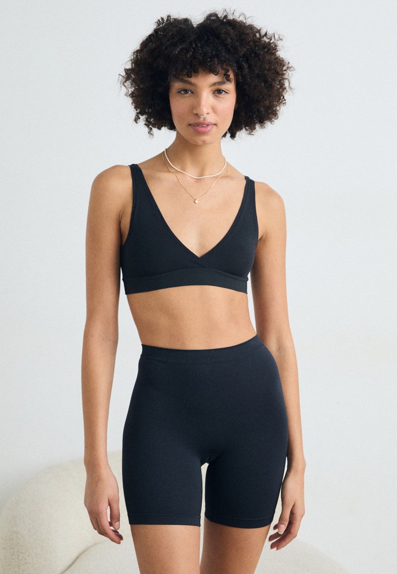 Musta ribbed-kangas bralette, jossa on syvä V-kaula-aukko ja leveät hihnat, yhdistettynä korkeavyötäröisiin ribbed-shorteihin, joilla on molemmilla tiivis malli.