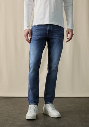 Dark blue denim jeans with a slim fit, featuring a slightly faded finish. Paired with white sneakers and a long-sleeve white top.